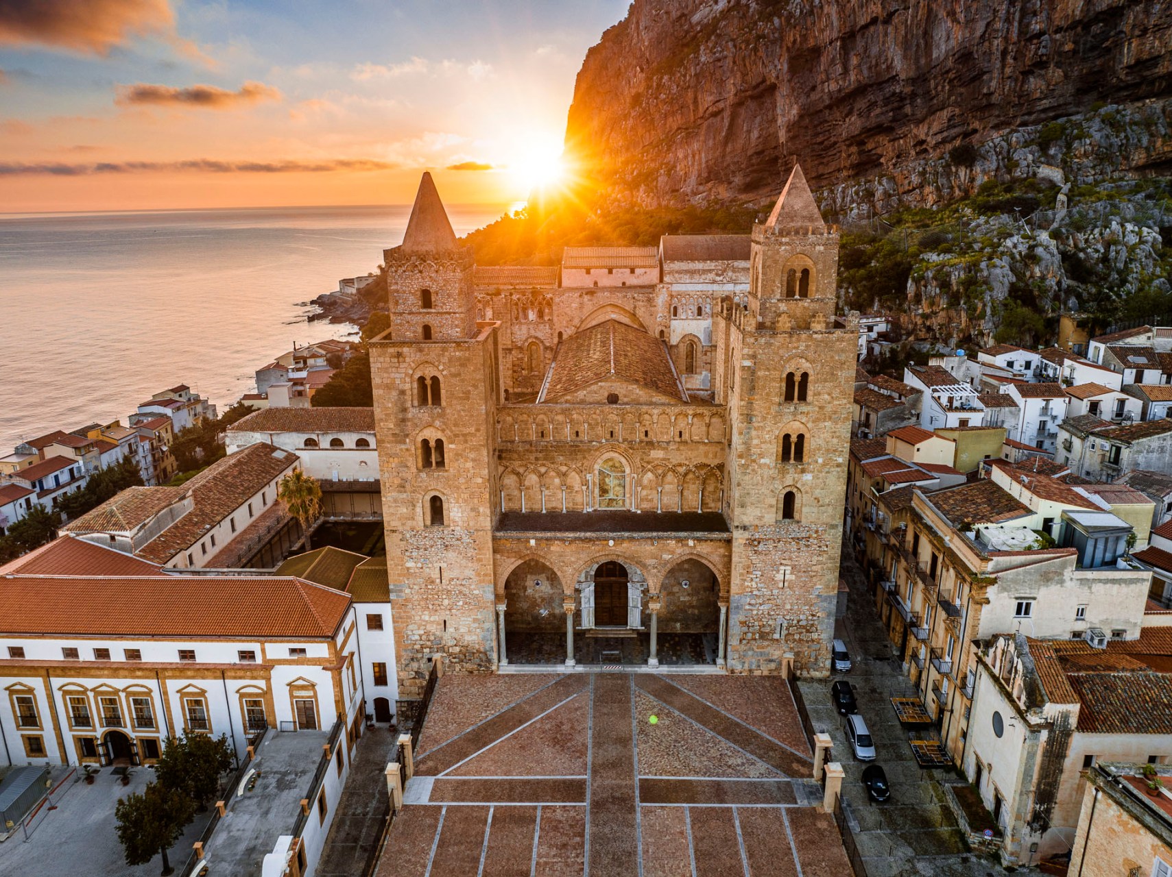 Fotografia aerea del Duomo di Cefalù all'alba con il sole che sorge dietro la Rocca e vista sul mare