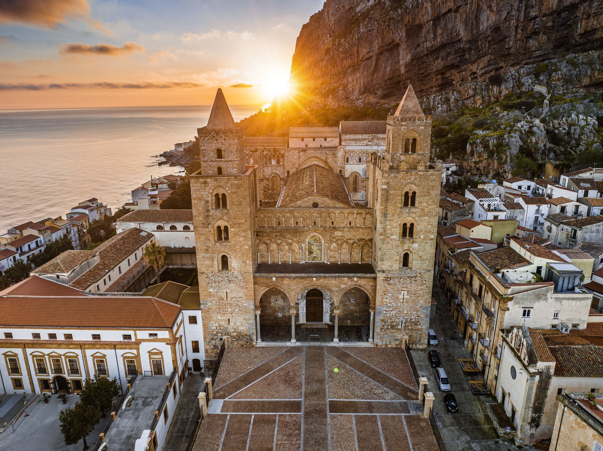 Fotografia aerea del Duomo di Cefalù all'alba con il sole che sorge dietro la Rocca e vista sul mare