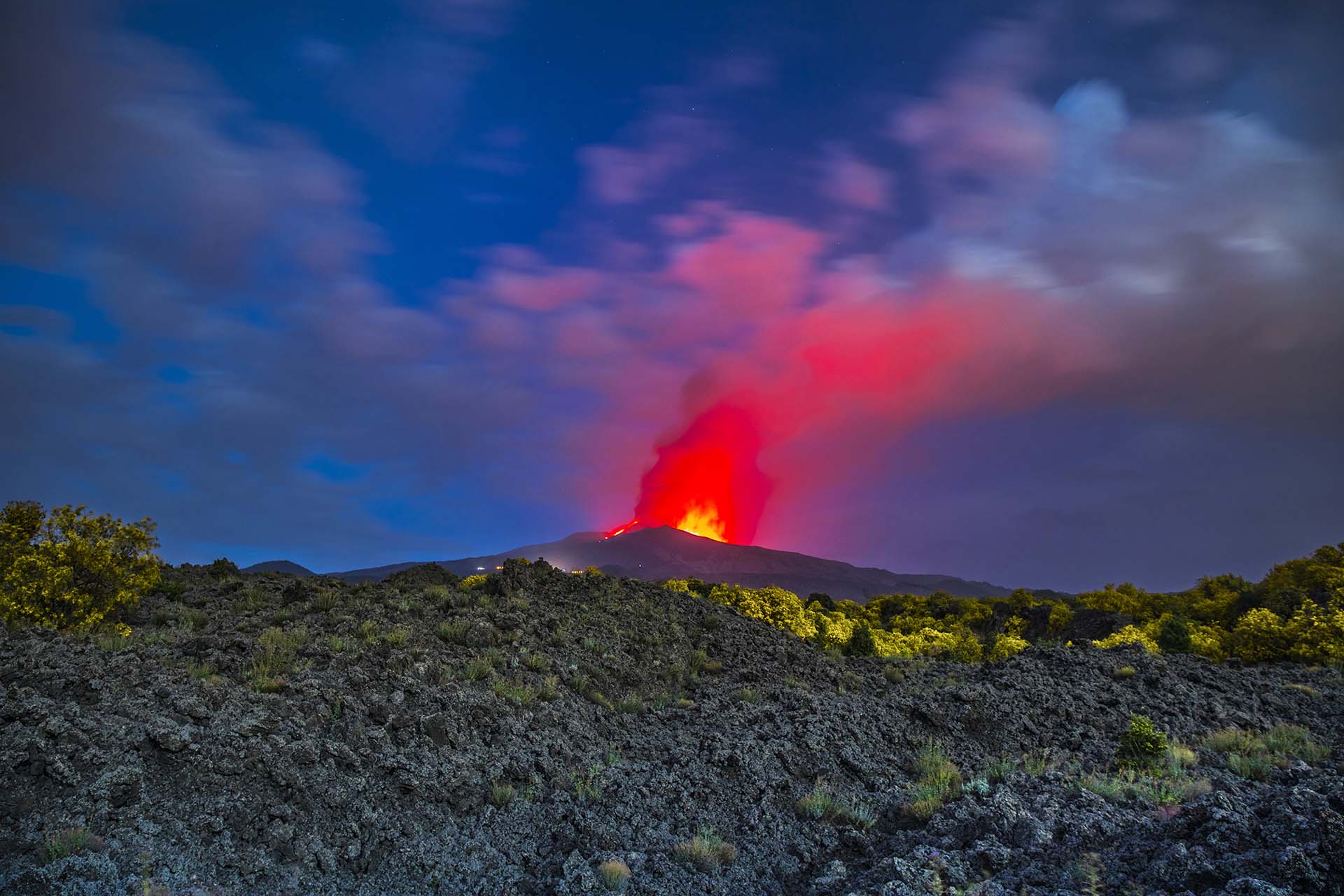 Etna, eruzione dai crateri sommitali vista dalle colate laviche di Serra Pizzuta piene di ginestre in fiore e illuminati dalla luna piena