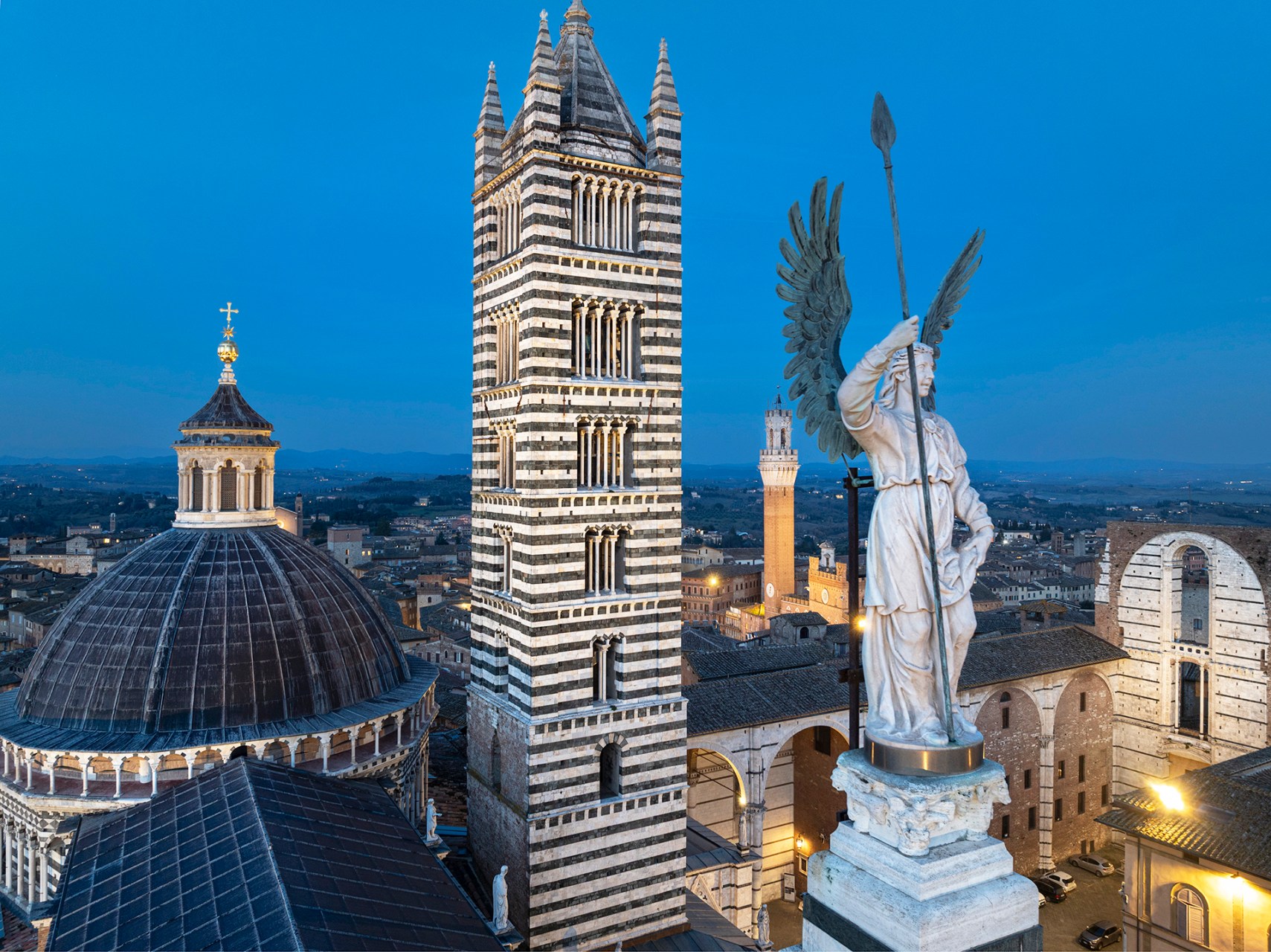 La statua in marmo di San Michele Arcangelo sulla cuspide centrale del Duomo di Siena con la cupola e il campanile.
