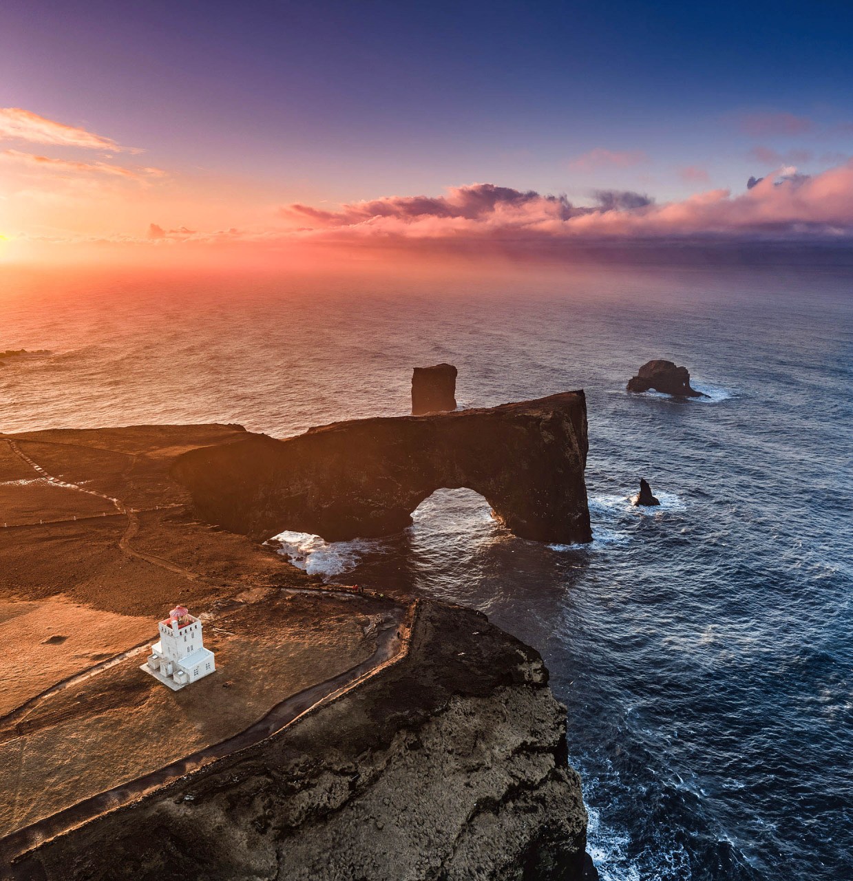Veduta aerea panoramica al tramonto di Dyrhólaey in Islanda, con il faro bianco, l'arco di roccia e l'oceano. Fotografia professionale drone di 101-zone.com