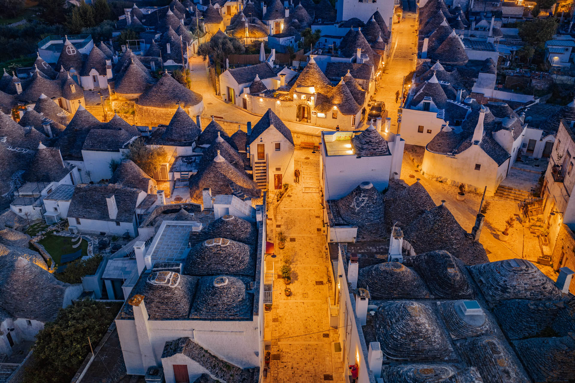 Inquadratura aerea notturna ravvicinata de I trulli di Alberobello (Puglia) illuminati. La foto cattura un vicolo accogliente e lastricato tra i coni di pietra, dove si intravedono alcune figure umane che passeggiano all'ora blu. La composizione esalta il contrasto tra il bianco candido delle pareti a calce e l'oro soffuso delle luci dei lampioncini, offrendo un'atmosfera autentica e immersiva del borgo UNESCO, firmata 101-zone.com