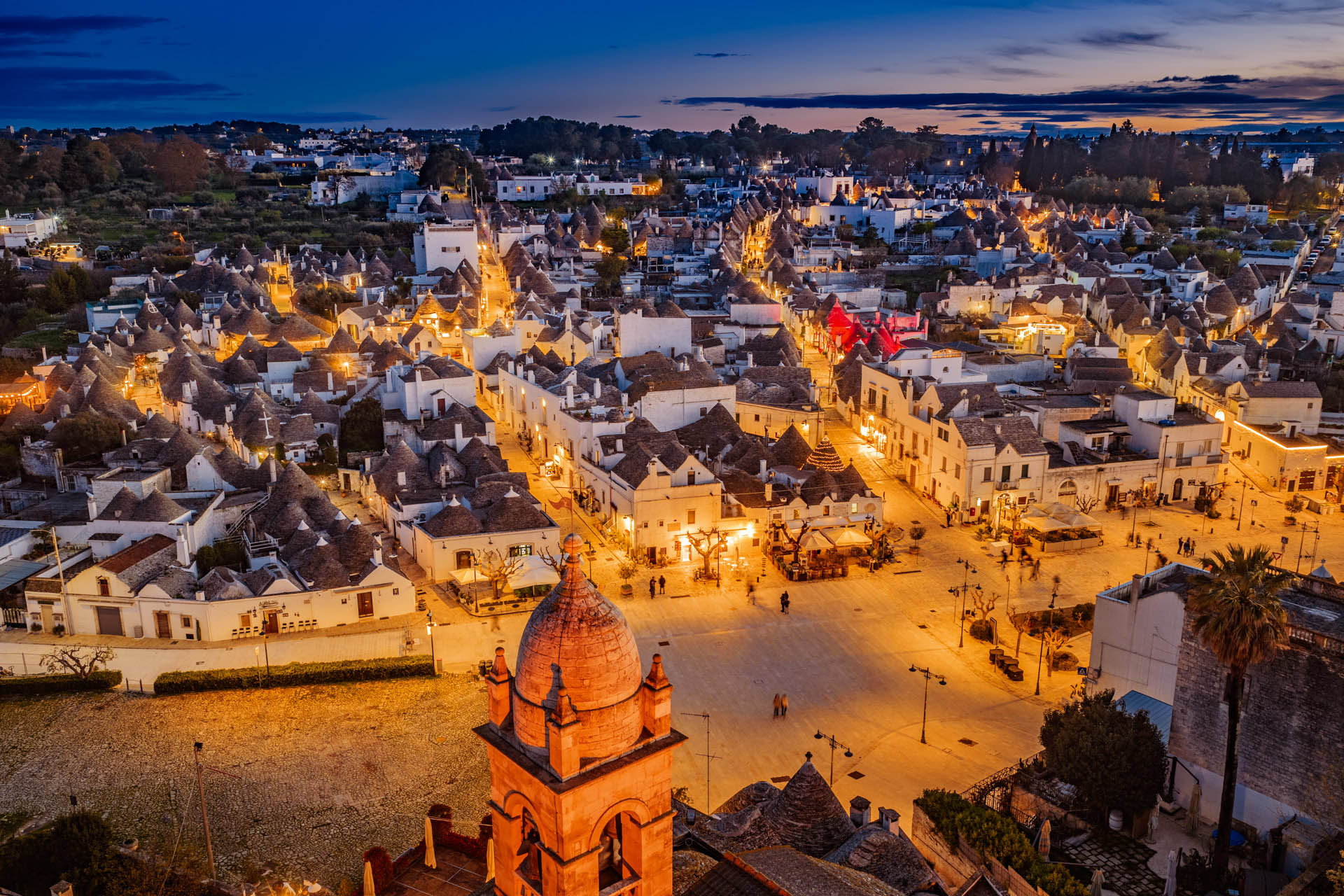 Veduta aerea panoramica notturna de I trulli di Alberobello (Puglia) all'ora blu. La fitta trama di coni in pietra illuminati si estende sotto un cielo crepuscolare blu profondo e caldo. In primo piano svetta la cupola barocca e il campanile della Chiesa Madre, creando un contrasto geometrico unico tra l'oro delle luci artificiali e il blu naturale