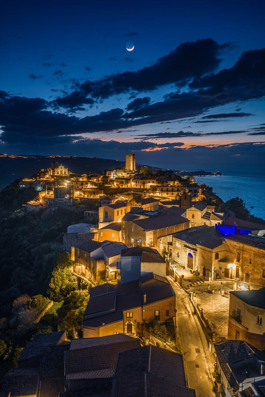 Veduta aerea notturna del centro storico di Piraino in Sicilia sotto la luna, con la torre saracena illuminata. Fotografia professionale drone di 101-zone.com