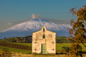 Chiesetta rurale con sfondo del Monte Etna innevato, immersa nel paesaggio della Piana di Catania.