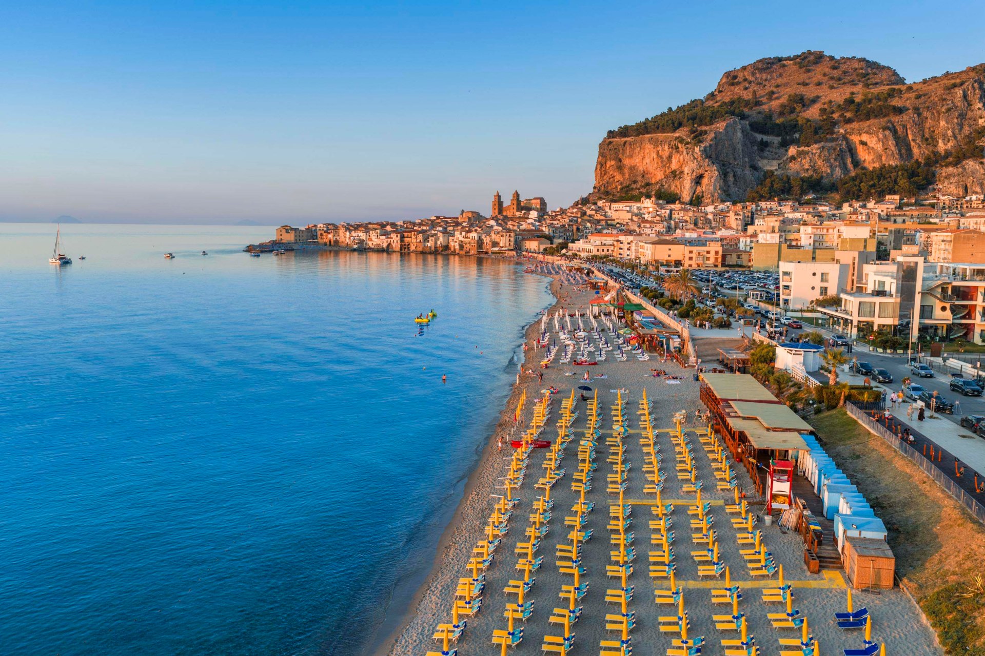 Fotografia aerea della spiaggia di Cefalù al tramonto con vista panoramica sul borgo, la Rocca e il mare calmo